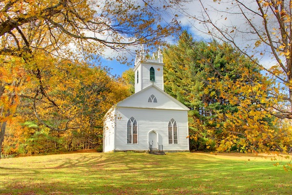 a white church with a steeple sitting in a green field, with trees with gold leaves around it.