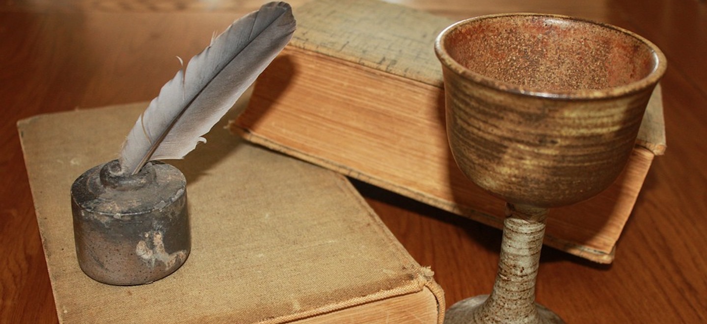 two old books, a chalice and an old style ink well with a quill in it sitting on a wooden table.