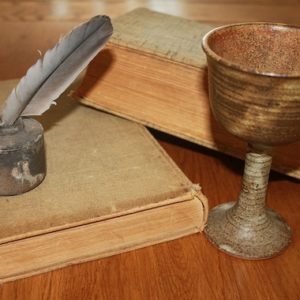 two old books, a chalice and an old style ink well with a quill in it sitting on a wooden table.