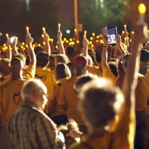 a march at night, with everyone facing away from the camera, holding electric candles up in the air, wearing gold t-shirts with hearts on the back