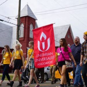 a group of people marching in a protest, most wearing gold t-shirts or hats that say side with love carrying a banner that has the UUA logo on it and says Unitarian Universalist Association.
