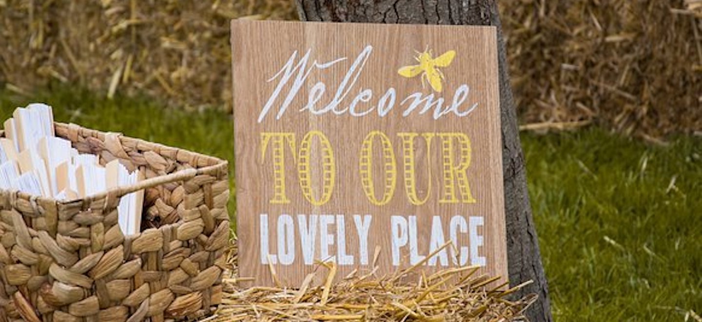 A board with Welcome to our lovely place painted on it, leaning on a tree, next to a woven basket with fans in it