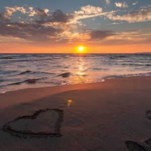 A sunrise over the ocean with a heart drawn in the sand and several footprints.