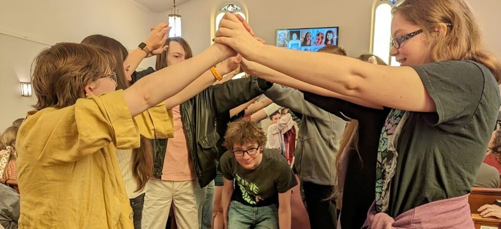 More than a dozen UU youth create a tunnel/bridge by reaching forward and holdng hands high so that bridging youth can travel through the created tunnel.