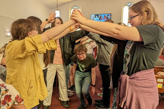 More than a dozen UU youth create a tunnel/bridge by reaching forward and holding hands high so that bridging youth can travel through the created tunnel.