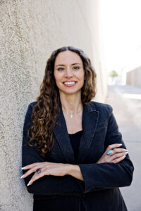 Lillian Drab-Braddick, wearing a black blazer, chalice necklace and long curly hair, in front of a textured wall with arms crossed.