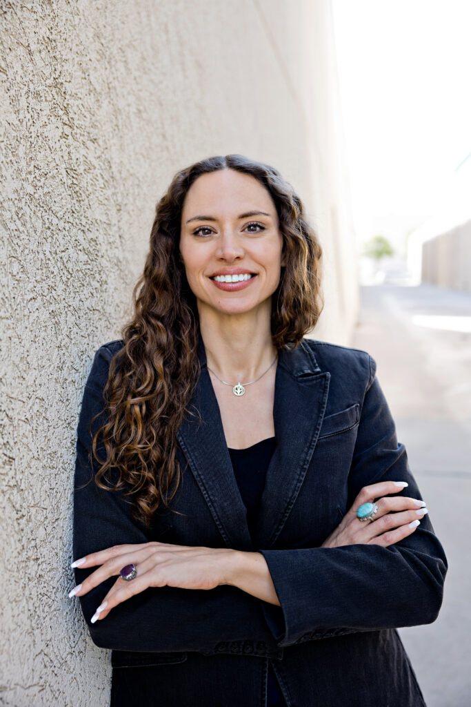 Lillian Drab-Braddick, wearing a black blazer, chalice necklace and long curly hair, in front of a textured wall with arms crossed.